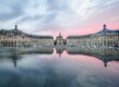 Place de la Bourse in Bordeaux, France, reflected in the Miroir d'eau water mirror at sunset, with pastel pink clouds and historic architecture.
