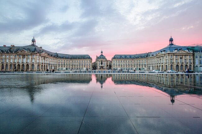 Place de la Bourse in Bordeaux, France, reflected in the Miroir d'eau water mirror at sunset, with pastel pink clouds and historic architecture.